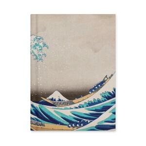 May include: A notebook featuring a print of Hokusai's "The Great Wave off Kanagawa." The design depicts a large ocean wave with a boat and Mount Fuji in the background. The color palette includes shades of blue, white, and beige.
