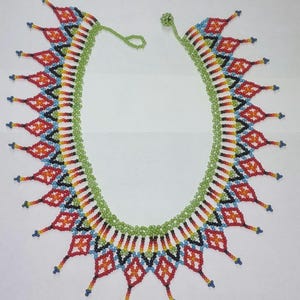 May include: A vibrant beaded necklace featuring a green, white, red, yellow, black, and blue design. The necklace has a green beaded edge and diamond-shaped patterns. The necklace is made of small, round beads.