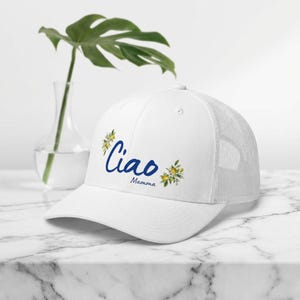 Może przedstawiać: Biała czapka typu trucker z napisem "Ciao Mamma" w niebieskim skrypcie, ozdobiona ilustracjami cytryn i kwiatów. Czapka ma siatkowy tył i zakrzywiony daszek.