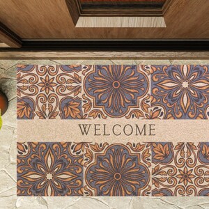 May include: A rectangular doormat featuring a repeating floral tile design in blue, orange, and beige. The word "WELCOME" is printed in black across the center. The mat is positioned in front of a wooden door.