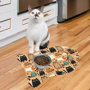 May include: A fish-shaped pet feeding mat featuring a repeating pattern of various cartoon cats in black, white, orange, and gray colors. A single silver-toned metal bowl filled with brown dry kibble sits in the center of the mat.