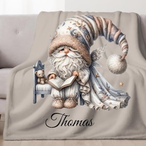 May include: A beige throw blanket featuring a watercolor illustration of a gnome reading a book. The gnome wears a tall hat and holds a book, with a teddy bear on a small chair. The name "Thomas" is written on the blanket.