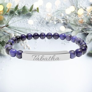 May include: A beaded bracelet with dark blue and purple spherical beads. The bracelet features a silver-colored rectangular bar engraved with the name "Tabatha". The bracelet is displayed on a white surface with decorative elements.