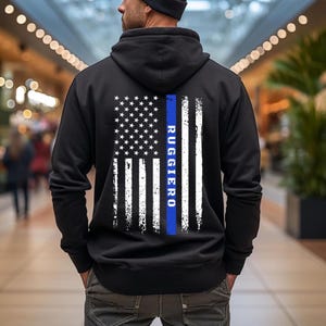 May include: Black hooded sweatshirt featuring a distressed American flag design on the back. The flag includes a blue stripe and the name "RUGGIERO" in blue vertical text. The sweatshirt is a casual garment.