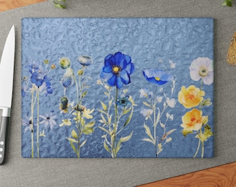 May include: A rectangular glass cutting board featuring a textured surface with a printed watercolor illustration of various wildflowers in shades of blue, yellow, and white against a mottled blue background, positioned next to a silver knife.