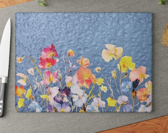 May include: A rectangular glass cutting board featuring a vibrant floral print of pink, yellow, orange, and blue wildflowers against a textured blue background that resembles water droplets. The board is positioned next to a silver kitchen knife.