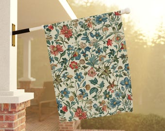May include: A floral garden flag with a cream background and a pattern of red, blue, and green flowers and leaves. The flag is hanging from a white pole attached to a house.