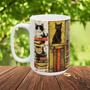 May include: A white ceramic mug featuring a wrap-around illustration of various cats perched on stacks of vintage books. The design includes a black and white kitten, a solid black cat, and a tabby cat, all set against a background of colorful book spines.