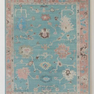 May include: A rectangular rug with a turquoise field and a pink border. The rug features a floral pattern in shades of pink, blue, and cream. It is displayed hanging from a gold-colored rod.