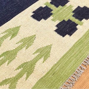 May include: Close-up of a woven rug with a geometric pattern. The rug features a navy blue corner, cream-coloured background, and green arrow and square designs. The edge has a green border and fringe.