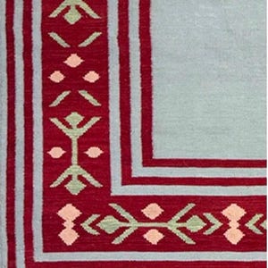 Könnte beinhalten: Rechteckiger Teppich mit hellblauem Feld und rotem Rand. Der Rand weist ein sich wiederholendes geometrisches Muster in Rot, Hellgrün und Pfirsich auf. Der Teppich hat einen Fransenbesatz am unteren Rand.