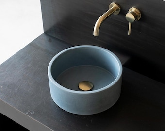May include: A blue concrete vessel sink with a gold drain stopper sits on a black metal countertop. A gold wall-mounted faucet is mounted on a black metal backsplash.