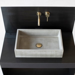 Op de afbeelding: Een betonnen wastafel met een messing kraan en afvoerstop, gemonteerd op een zwart metalen aanrecht.