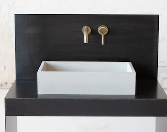 Op de afbeelding: Een rechthoekige betonnen wastafel in wit met een gouden kraan en hendel gemonteerd op een zwarte muur. De wastafel staat op een zwart metalen onderstel met een zwart aanrecht.
