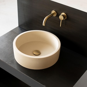 Könnte beinhalten: Ein rundes, cremefarbenes Keramikwaschbecken mit einem goldenen Abflussstopfen sitzt auf einer schwarzen Arbeitsplatte. Ein goldener Wasserhahn und ein goldener Griff sind an einer schwarzen Wand hinter dem Waschbecken montiert.