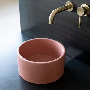 May include: A round, pink concrete vessel sink with a gold faucet mounted on a black wall.