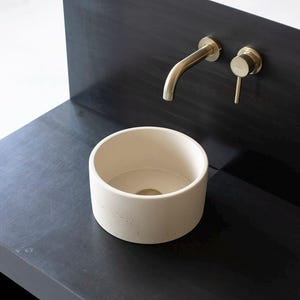 May include: A modern bathroom sink setup featuring a round, off-white basin set into a dark gray countertop. The faucet and handle are gold-toned, adding a touch of elegance. The design is minimalist and contemporary.