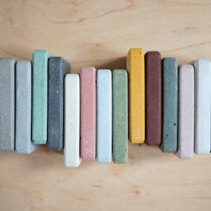 May include: A collection of colorful, rectangular coasters in various shades, including gray, green, yellow, and pink. The coasters are arranged vertically against a light wood grain background, showcasing their textured surfaces.