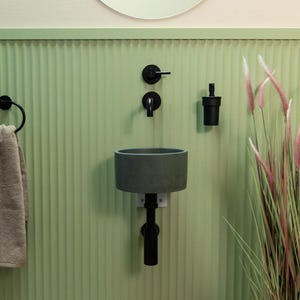Op de afbeelding: Een moderne badkameropstelling met een grijze cilindrische wastafel, zwarte kraan en zeepdispenser tegen een lichtgroene, verticaal geribbelde muur. Een handdoekring en decoratieve planten dragen bij aan het minimalistische ontwerp.