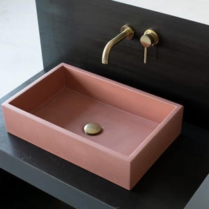May include: A rectangular, pink concrete sink with a brass faucet and drain stopper. The sink is mounted on a black countertop.