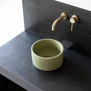 Op de afbeelding: Een ronde, groene keramische wastafel op een zwart aanrecht. Een gouden kraan is gemonteerd aan de muur achter de wastafel.