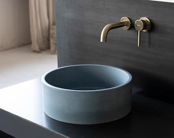 May include: A round, light blue concrete vessel sink with a gold faucet and handle mounted on a black wall.