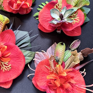 Può includere: Tre fermagli per capelli floreali con petali di orchidea rosso vivo, accenti di dionaea muscipula verde e piccoli elementi di piante carnivore. Un fermaglio include un piccolo insetto artificiale su una foglia verde. Sfondo di colore scuro.