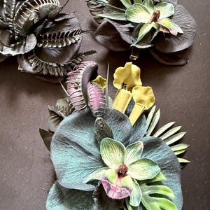 May include: Three hair clips featuring artificial botanical arrangements. Each clip includes dark-toned orchid petals, green fern-like foliage, and purple-tinted Venus flytrap structures. One clip also features yellow pitcher plant-like elements.