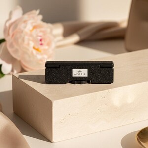 May include: A rectangular black object with a textured, speckled finish and a hinged lid. A white rectangular label is centered on the front, featuring the text AVORIE in black capital letters with a stylized line symbol above the brand name.