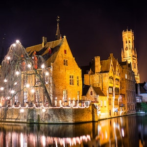 Puede incluir: Vista nocturna de edificios iluminados a lo largo de un canal. Los edificios son de ladrillo y tienen un brillo cálido y dorado de las luces. Una alta torre de campanas es visible en el fondo, y el agua refleja las luces.