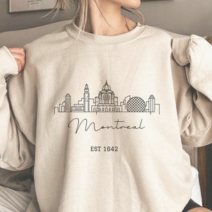 May include: A beige sweatshirt with a black outline of the Montreal skyline and the text "Montreal EST 1642".