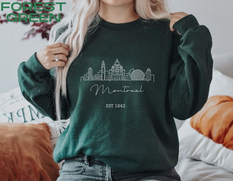 May include: Forest green sweatshirt with a white outline of the Montreal skyline, including the words "Montreal" and "EST 1642".