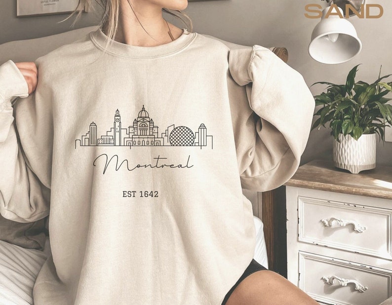 May include: A beige sweatshirt with a black outline of the Montreal skyline and the text "Montreal EST 1642".