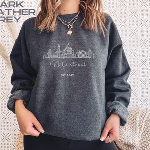 May include: Dark heather grey sweatshirt with a white outline of the Montreal skyline and the text "Montreal Est 1642".