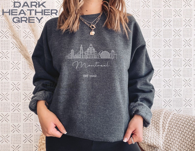 May include: Dark heather grey sweatshirt with a white outline of the Montreal skyline and the text "Montreal Est 1642".