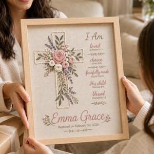 May include: A framed embroidery art piece on light fabric featuring a central cross adorned with pink and white floral stitching. The text reads: I Am loved, chosen, fearfully made, His child, blessed, Emma Grace, and Baptized on February 1st, 2026.