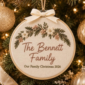 May include: A round wooden embroidery hoop ornament featuring cream-colored fabric with an embroidered floral wreath in green, white, and pink. The text reads The Bennett Family and Our Family Christmas 2026. A cream ribbon bow sits at the top.