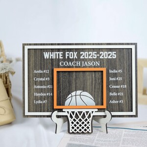 May include: A wooden basketball-themed sign with the text "WHITE FOX 2025-2025 COACH JASON". The sign features a basketball hoop design with a basketball and a list of names and numbers. The sign is supported by two white stands.