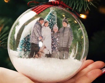 May include: A clear glass Christmas bauble with a red and black checked ribbon. Inside the bauble is a photo of a family of four standing in front of a small Christmas tree. The bauble is filled with white faux snow.
