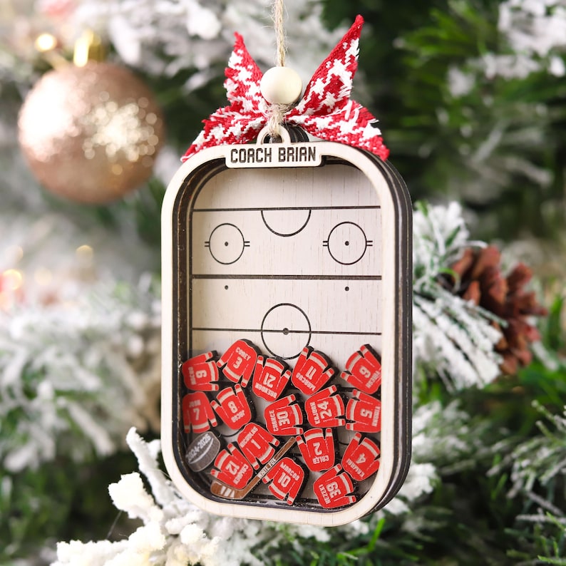 May include: A wooden hockey-themed Christmas ornament. The ornament features a hockey rink design with miniature red jersey-shaped pieces inside. The top of the ornament has the text "COACH BRIAN" and is adorned with a red and white bow.