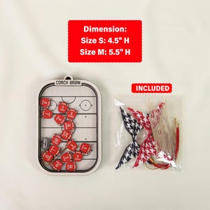 May include: A hockey-themed ornament with a wooden frame, featuring a miniature hockey rink and red jersey pieces. The ornament is 11.4 cm or 14 cm high. Included are decorative bow ties and hanging strings.