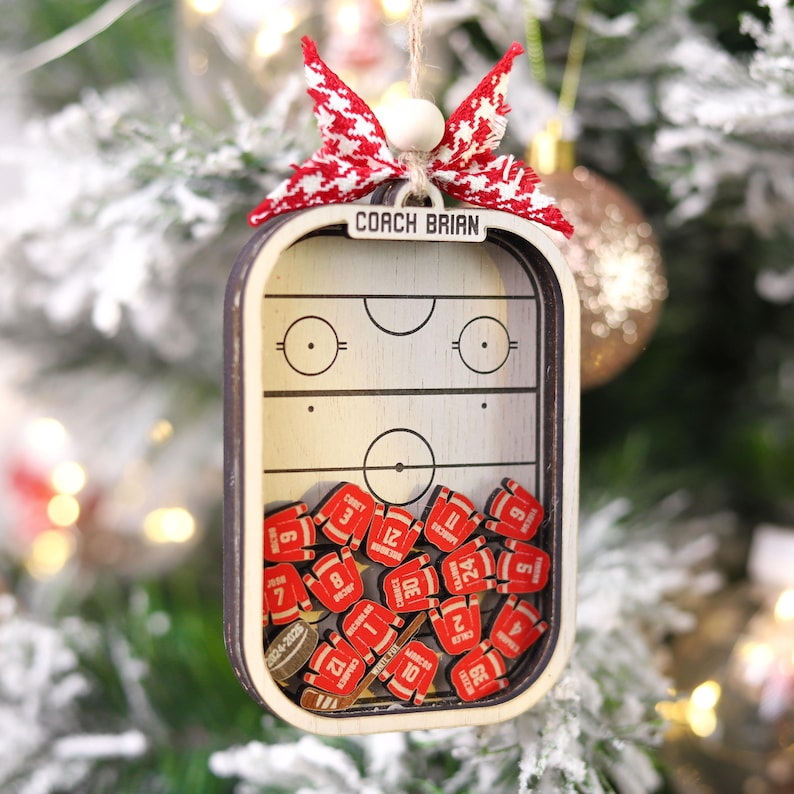 May include: Wooden hockey-themed Christmas ornament with the text "COACH BRIAN." The ornament features a miniature hockey rink design and small red jersey-shaped pieces with numbers. A red and white bow tops the ornament, which hangs from a string.