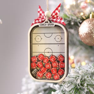 May include: Wooden hockey-themed Christmas ornament with a red and white bow. The ornament features a hockey rink design with miniature red jersey-shaped pieces inside. The text "COACH BRIAN" is displayed at the top.