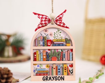 May include: Wooden bookshelf ornament with a red and white houndstooth bow. The ornament features a miniature bookshelf design filled with colorful books, plants, and a mug that says "I LOVE". The name "GRAYSON" is printed at the bottom.