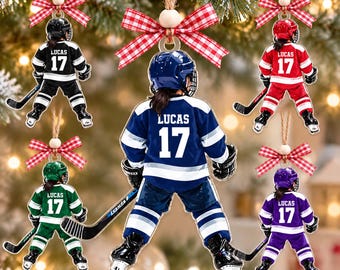 May include: Five flat hanging ornaments shaped like hockey players wearing helmets, jerseys, and skates. Each player has the name LUCAS and the number 17 printed on the back. The jerseys are black, red, blue, green, and purple with plaid bows.