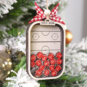 May include: Wooden hockey-themed Christmas ornament. The ornament features a miniature hockey rink design with the text "COACH BRIAN" at the top. Inside are miniature red hockey jerseys with numbers. A red and white bow and a wooden bead are at the top.