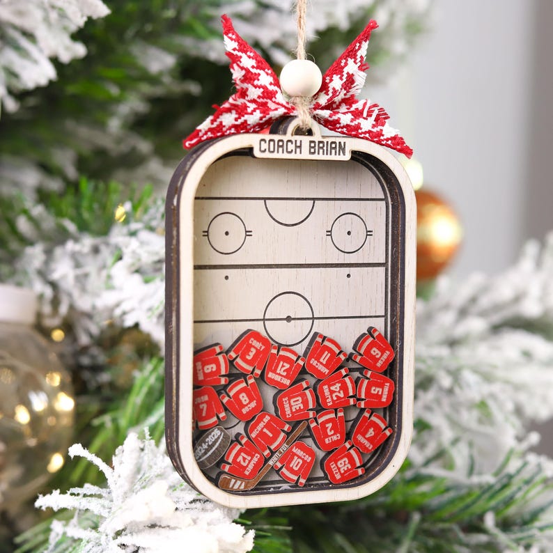 May include: Wooden hockey-themed Christmas ornament. The ornament features a miniature hockey rink design with the text "COACH BRIAN" at the top. Inside are miniature red hockey jerseys with numbers. A red and white bow and a wooden bead are at the top.