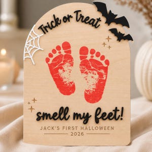 May include: An arched light wood plaque featuring red baby footprints in the center. The text reads Trick or Treat and smell my feet! in black script. It includes a white spiderweb, black bats, and small star cutouts, with JACK'S FIRST HALLOWEEN 2026.