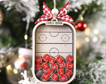 May include: Wooden hockey-themed Christmas ornament. The ornament features a hockey rink design with the text "COACH BRIAN" and small red jersey-shaped pieces with numbers. It is decorated with a red and white bow.