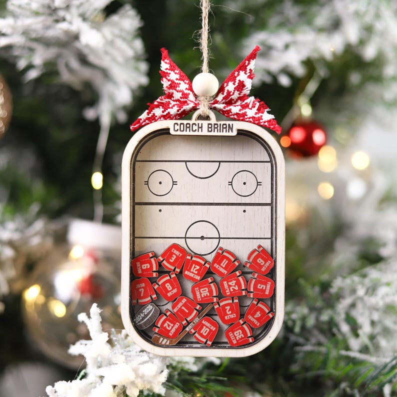 May include: Wooden hockey-themed Christmas ornament. The ornament features a hockey rink design with the text "COACH BRIAN" and small red jersey-shaped pieces with numbers. It is decorated with a red and white bow.
